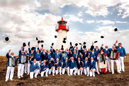 Eine Gruppe eines Shantychors in blauen Jacken und weißen Hosen wirft schwarze Mützen in die Luft vor einem Leuchtturm.