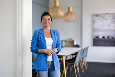 Eine Frau mit braunen Haaren und freundlichem Ausdruck steht in einem modernen Besprechungsraum. Sie trägt einen blauen Blazer über einem weißen Oberteil und Jeans. Im Hintergrund sind ein langer Tisch, Stühle und dekorative Hängeleuchten zu sehen.