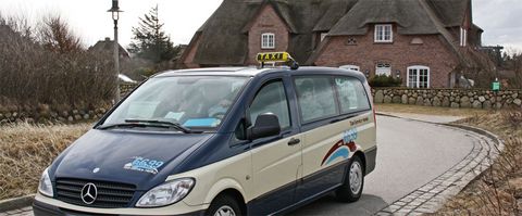 Ein Mercedes-Taxi-Van von „Taxi-Service Henke“ fährt auf einer gepflasterten Straße durch eine ländliche Gegend mit traditionellen Reetdachhäusern auf Sylt.