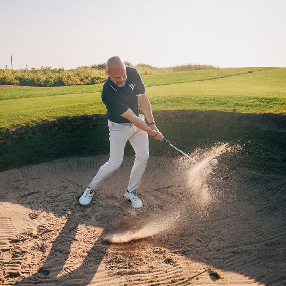 Golfer schlägt Ball aus Sandbunker, Sand fliegt auf. 