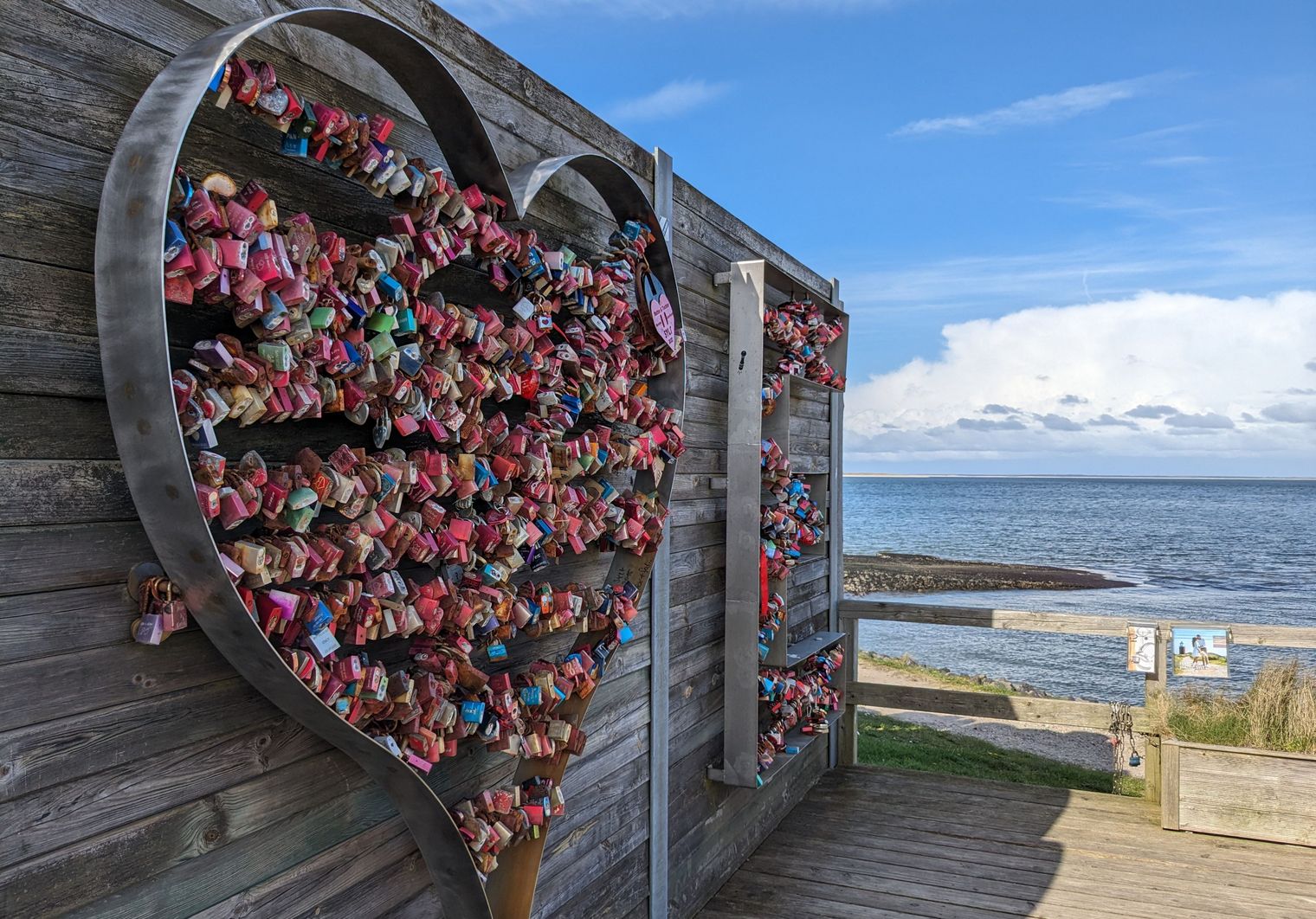 Metallherz voller bunter Liebesschlösser an einer Holzwand vor Küstenlandschaft und ruhigem Meer.