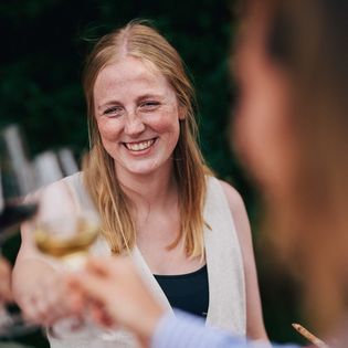 Lächelnde Frau stößt mit anderen Gästen bei einem Glas Wein in einer sommerlichen Umgebung an