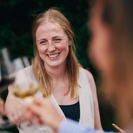 Lächelnde Frau stößt mit anderen Gästen bei einem Glas Wein in einer sommerlichen Umgebung an