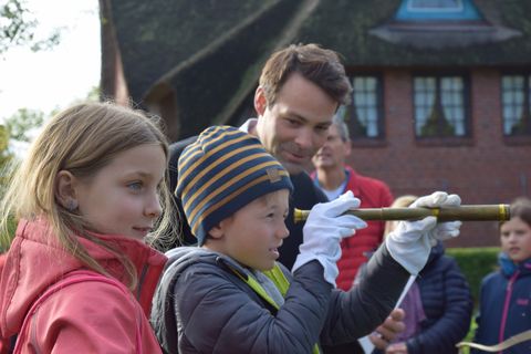 Ein Junge mit gestreifter Mütze schaut durch ein historisches Fernrohr, während ein Mann ihm dabei lächelnd über die Schulter schaut. Neben dem Jungen steht ein Mädchen, das ebenfalls interessiert zusieht. Im Hintergrund sind weitere Personen und ein traditionelles Reetdachhaus zu sehen.