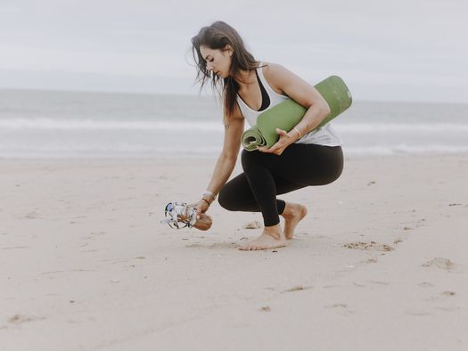 Eine Frau sammelt achtsam Müll am Strand ein, während sie eine zusammengerollte Yogamatte unter dem Arm trägt.