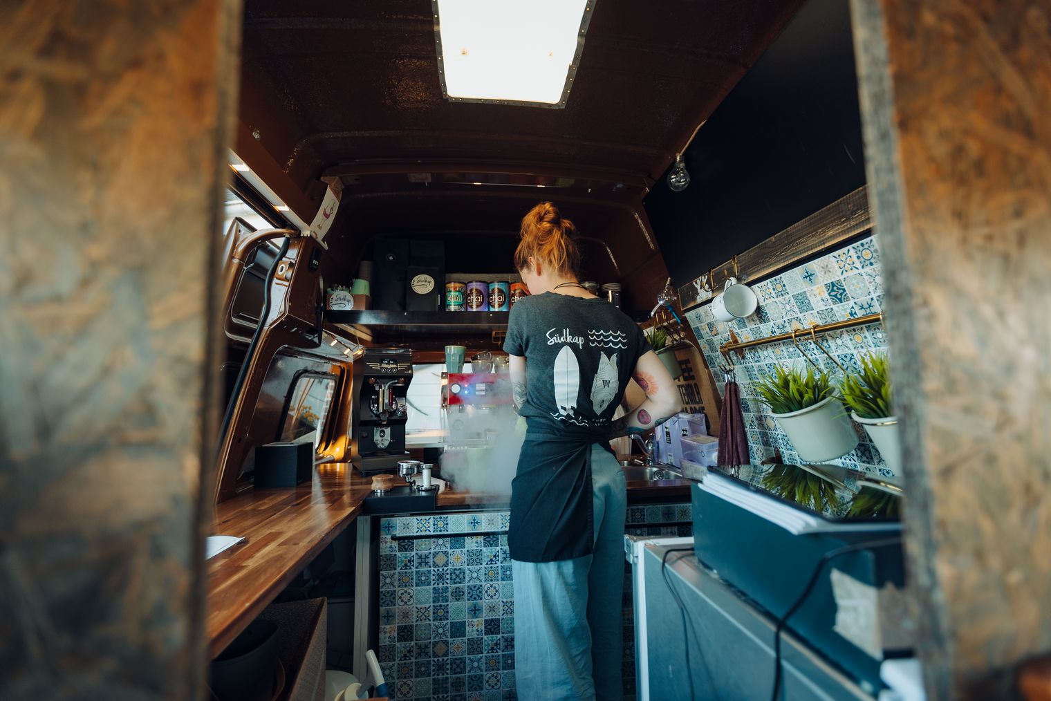 Barista bereitet Kaffee in einem stilvollen Kaffeebus mit Holzelementen und Fliesenoptik zu.