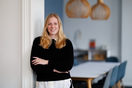 Eine Frau mit langen blonden Haaren steht lächelnd an einer weißen Wand in einem modernen Büro. Sie trägt einen schwarzen Pullover und hat die Arme vor der Brust verschränkt. Im Hintergrund sind zwei große hölzerne Designer-Lampen und ein Besprechungstisch mit Stühlen zu sehen.