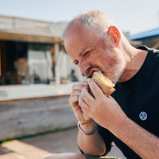 Mann isst Snack vor rustikalem Gebäude bei Sonnenschein.