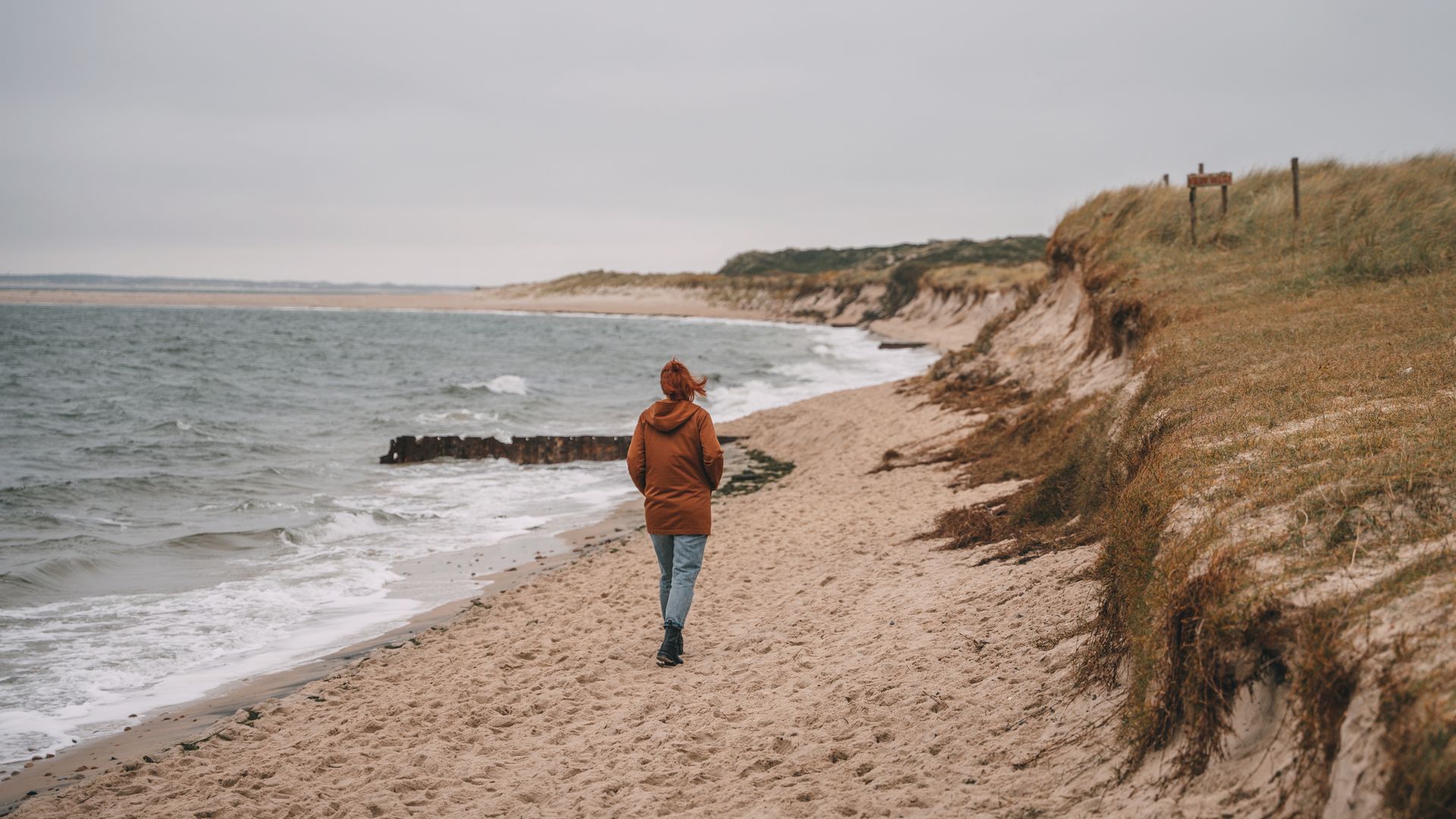 Person mit brauner Jacke spaziert an einem sandigen Strand entlang der Dünen.