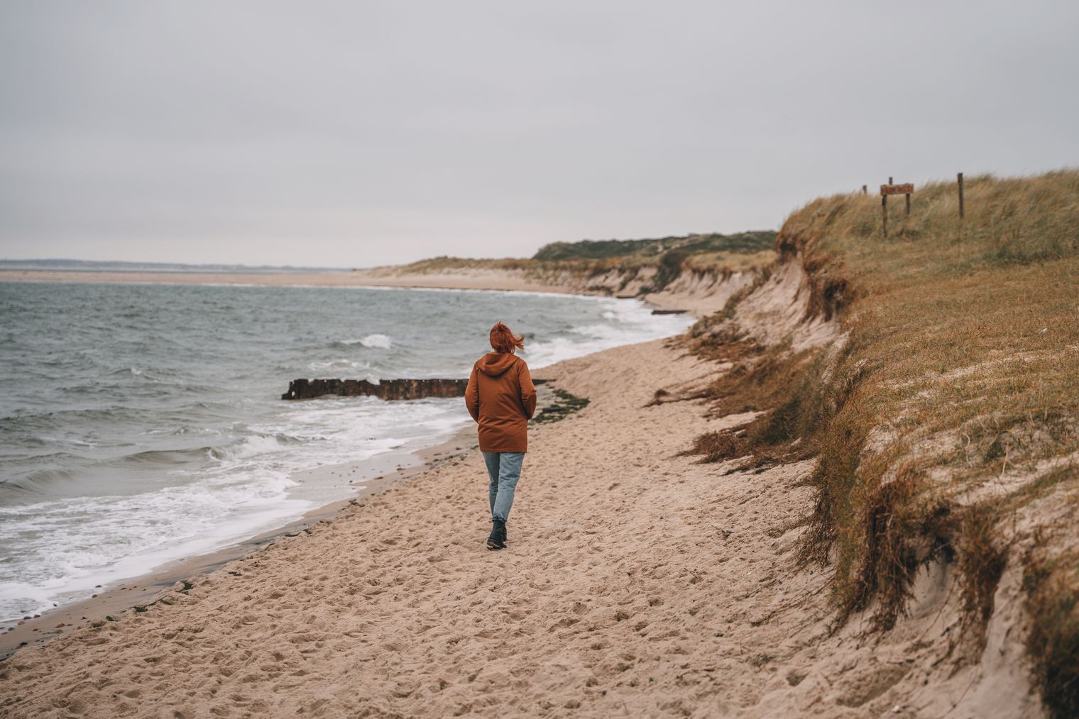 Person mit brauner Jacke spaziert an einem sandigen Strand entlang der Dünen.