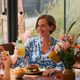 Eine Frau in einem blau-weißen Kleid sitzt im Twisters Beach Dinner in Wenningstedt auf Sylt, hält ein Cocktailglas und lächelt an einem Tisch mit Burger, Pommes und Blumen.