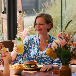 Eine Frau in einem blau-weißen Kleid sitzt im Twisters Beach Dinner in Wenningstedt auf Sylt, hält ein Cocktailglas und lächelt an einem Tisch mit Burger, Pommes und Blumen.