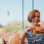 Lachende Frau isst einen Burger in einem Restaurant mit Ausblick auf Gräser und blauen Himmel.