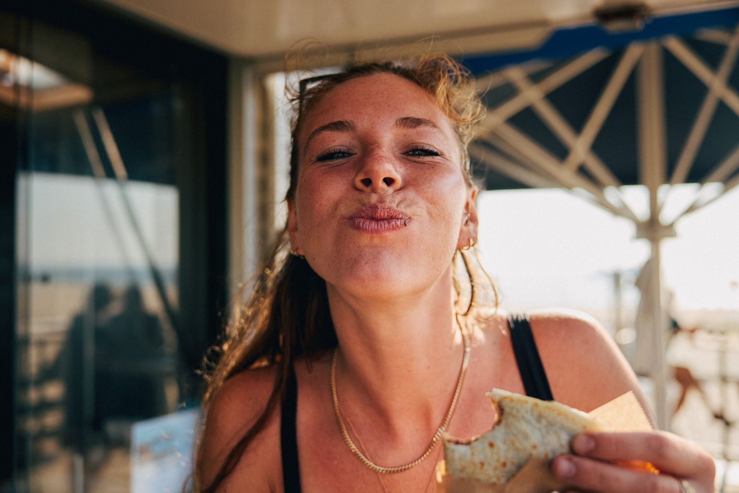 Frau in Sommerkleidung genießt Snack vor Sonnenschirm und Strand.