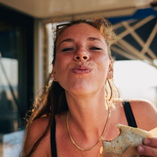 Frau in Sommerkleidung genießt Snack vor Sonnenschirm und Strand.