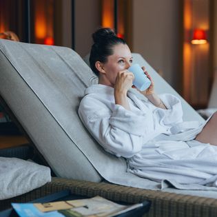 Eine Frau in einem weißen Bademantel liegt entspannt auf einer Liege und genießt ein warmes Getränk.