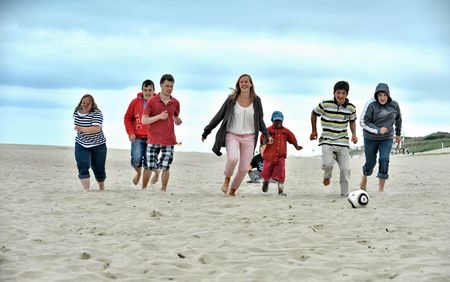 Eine Gruppe junger Menschen läuft fröhlich über den Sandstrand, während sie einem Fußball nachjagen. Der Himmel ist bewölkt, und die Stimmung ist lebendig.