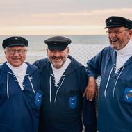 Drei Männer in maritimen Uniformen und Mützen lächeln vor einem Strandkorb, mit Meer und Sonnenuntergang im Hintergrund.