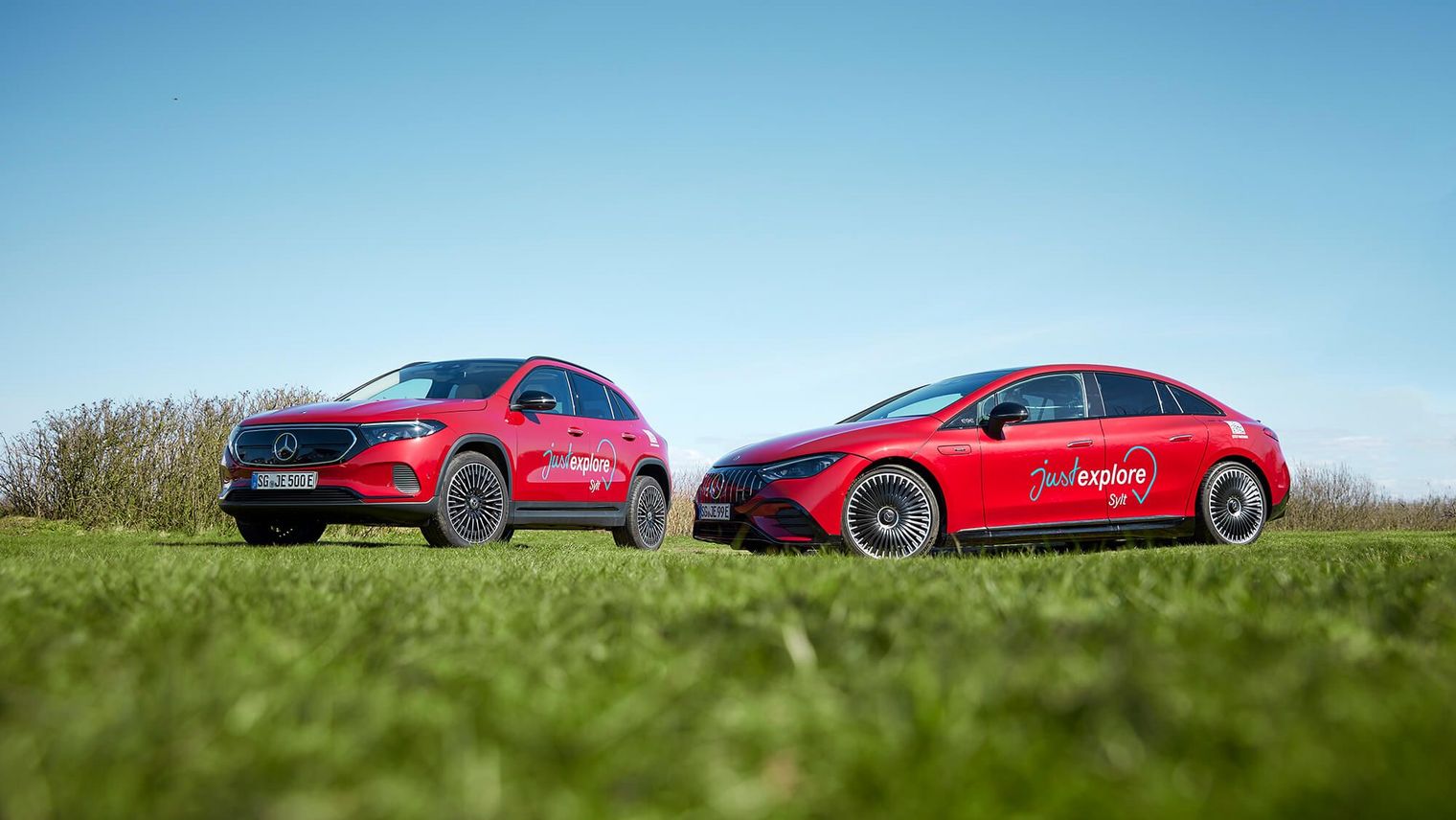 Zwei rote Mercedes-Fahrzeuge mit dem Schriftzug "just explore Sylt" stehen auf einer Wiese unter einem blauen Himmel. Links ein SUV, rechts eine Limousine.