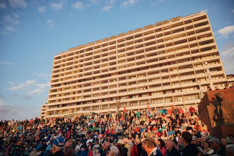  Menschenmenge sitzt auf den Stufen vor der Musikmuschel in Westerland vor einem großen Hochhaus in warmem Sonnenlicht. 