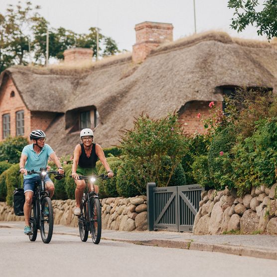 Ein Mann und eine Frau fahren nebeneinander auf modernen E-Bikes durch eine ruhige Wohngegend auf Sylt. 