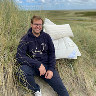 Mann mit Brille und Kapuzenpullover sitzt in den Dünen neben einer Decke.