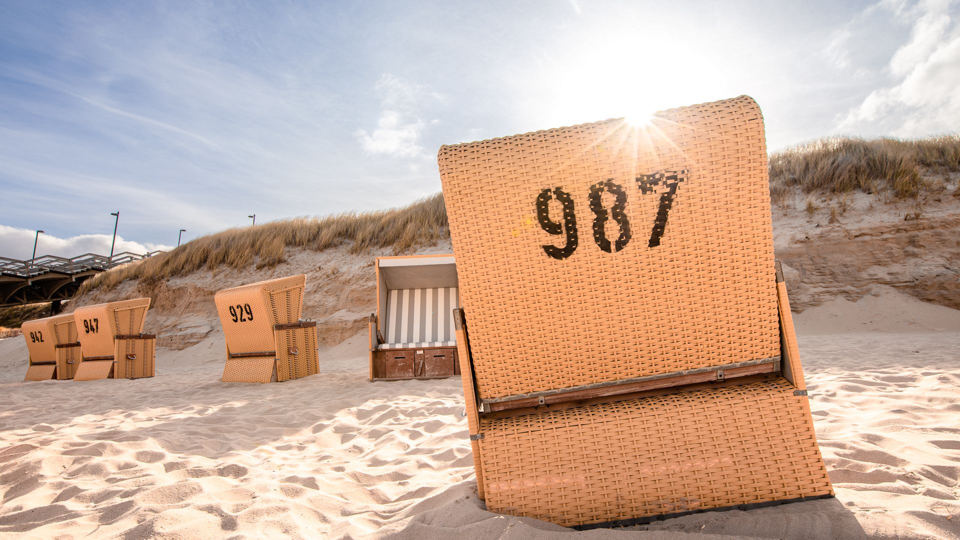 Strandkorb mit Nummer "987" auf Sandstrand vor Dünen und blauem Himmel.