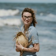 Frau mit Strohhut lächelt am Strand, Wind weht durch ihr Haar.