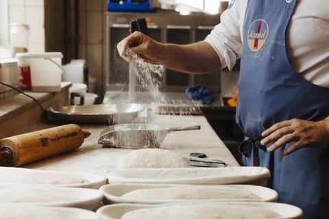 Ein Bäcker in blauer Schürze streut Mehl über einen Teiglaib auf einer Arbeitsfläche. Neben ihm liegen mehrere geformte Teiglinge in Körbchen, bereit zum Backen. Im Hintergrund sind verschiedene Küchenutensilien und Behälter zu sehen.