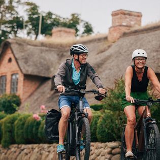 Zwei Personen auf E-Bikes fahren durch Keitum auf Sylt. Im Hintergrund sind Reetdachhäuser sichtbar.