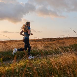 Eine Frau läuft bei Sonnenaufgang durch eine Wiesenlandschaft, während Gräser im Vordergrund wehen. 