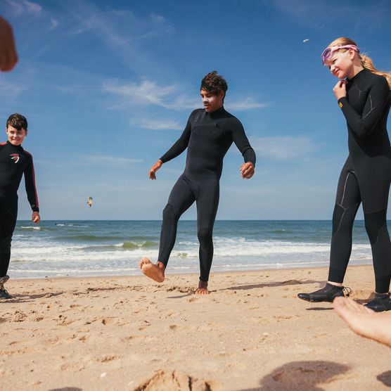 Jugendliche in Neoprenanzügen spielen barfuß am Strand nahe dem Meer.