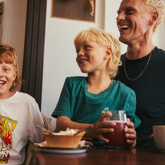 Ein Mann und zwei Kinder lachen gemeinsam, während sie an einem Tisch sitzen. Die Kinder trinken aus Gläsern, auf dem Tisch steht eine Waffel mit Sahne.