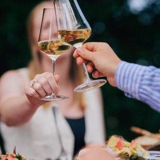 Lächelnde Frau stößt mit anderen Gästen bei einem Glas Wein in einer sommerlichen Umgebung an.