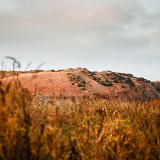 Rote Klippe mit herbstlichen Gräsern im Vordergrund und bewölktem Himmel.