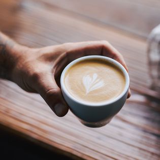 Hand hält Tasse Cappuccino mit Latte-Art-Muster vor Holztisch.