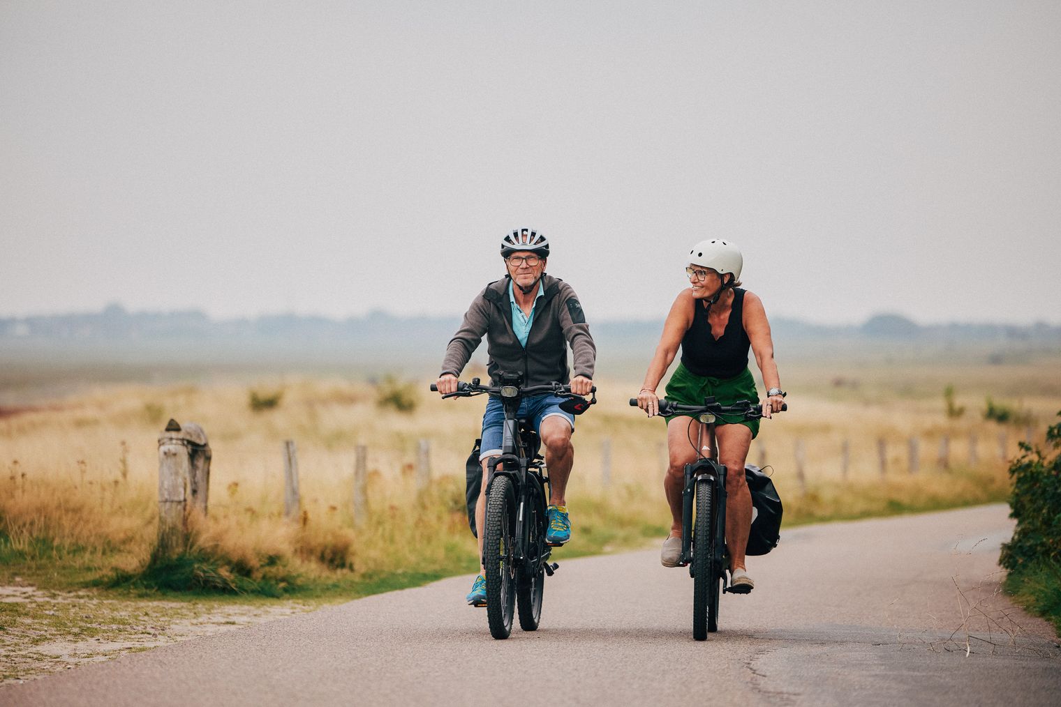 Zwei Personen auf E-Bikes fahren lächelnd durch eine offene, grasbewachsene Landschaft.