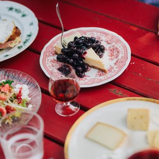 Teller mit Käsekuchen, Blaubeeren, Salat und Käse sowie ein Glas Rotwein auf rotem Tisch.