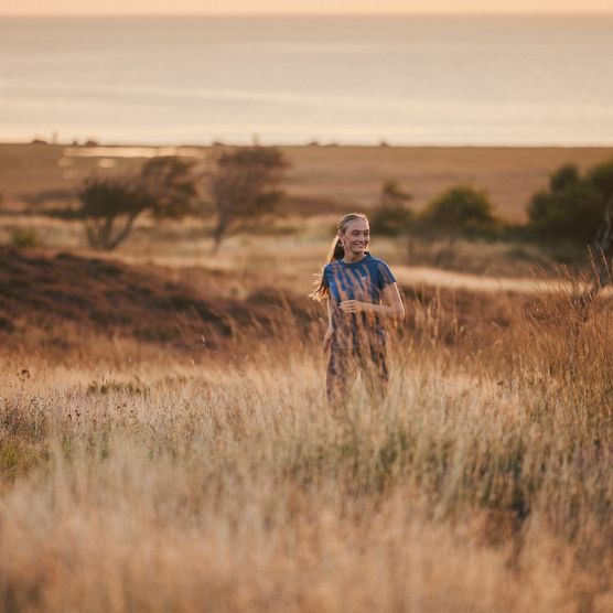 Eine Frau joggt lächelnd durch hohe Gräser bei Sonnenaufgang, mit Meer und weiten Feldern im Hintergrund. 