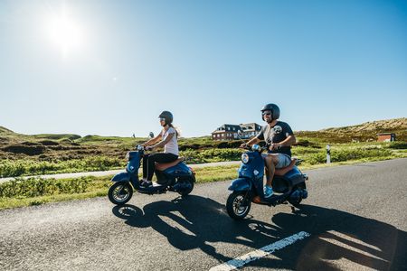 Ein Mann und eine Frau fahren mit blauen E-Rollern über die Insel Sylt. 