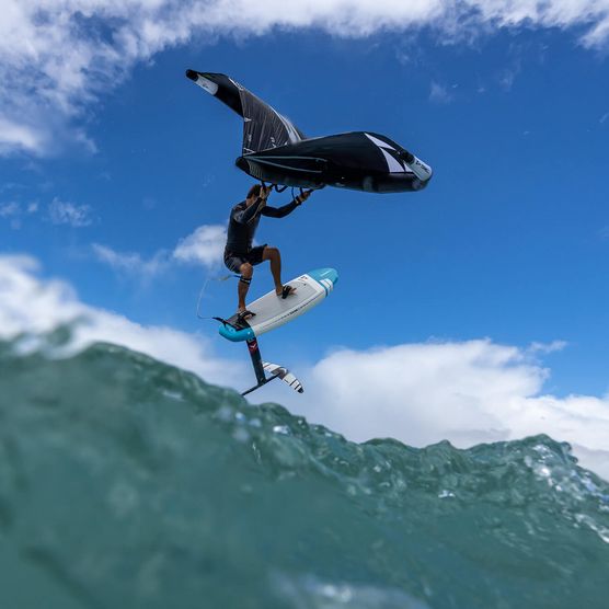 Ein Wingfoiler auf einem Foilboard hebt über den Wellen ab, mit einem strahlend blauen Himmel und weißen Wolken im Hintergrund.