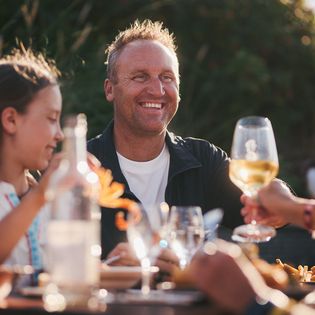 Eine Frau hebt lächelnd ein Glas Weißwein, während sie mit einer anderen Person im Freien anstößt. Daneben sitzt ein Kind.