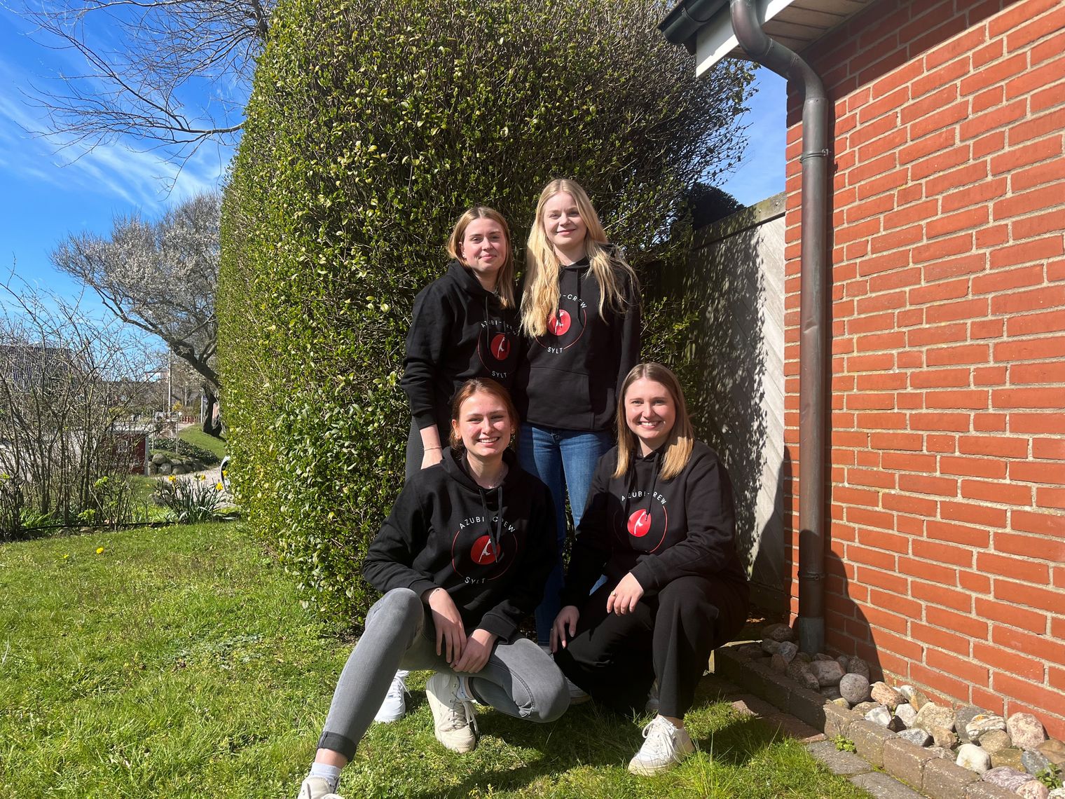 Vier junge Frauen, Mitglieder der Azubi-Crew Sylt 2024, posieren lächelnd für ein Gruppenfoto. Sie tragen schwarze Hoodies mit einem roten Logo und stehen in einem Garten vor einer Hecke neben einer Backsteinwand.