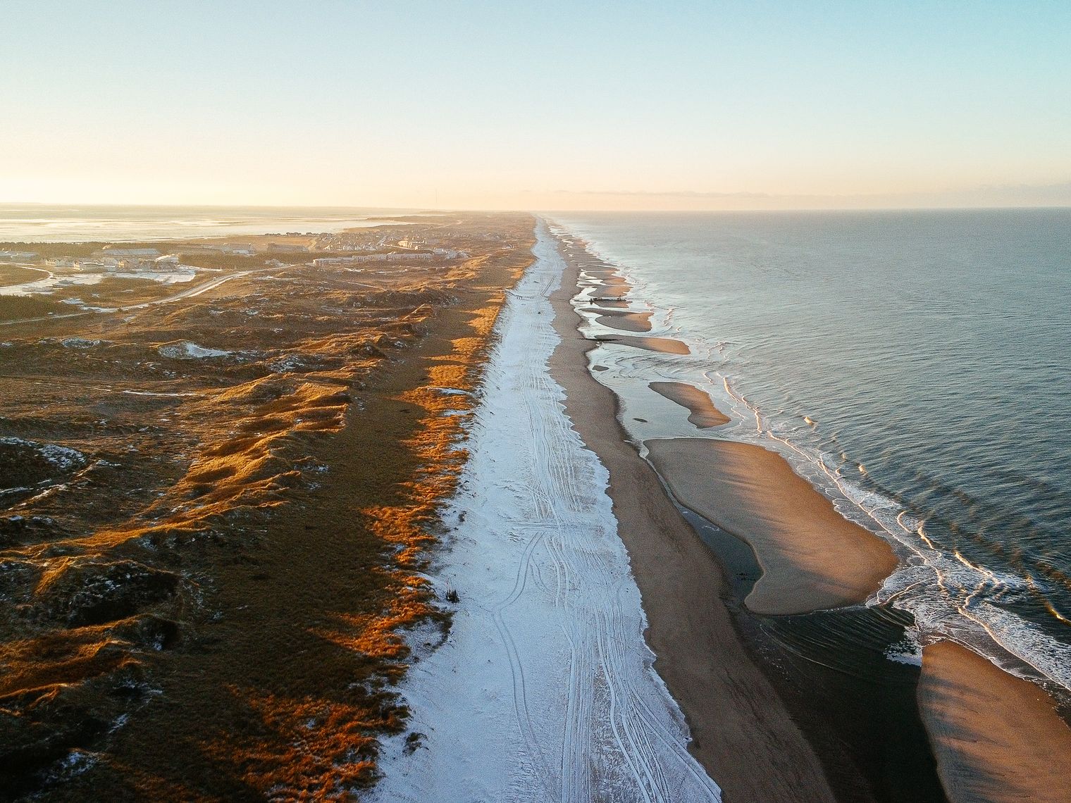 Luftaufnahme eines vereisten Strands auf Sylt mit Dünen und Siedlungen links sowie Meer und sanften Wellen rechts. Die Szene ist in warmes Sonnenlicht getaucht.