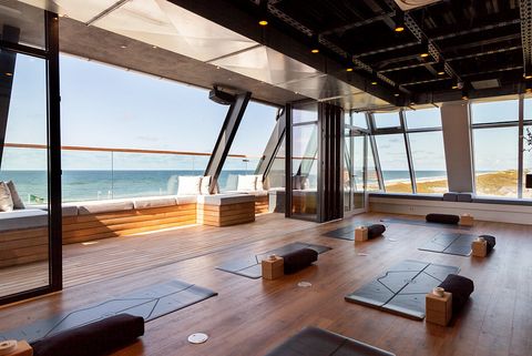 Ein moderner Yoga-Raum mit großen Glasfenstern, durch die man einen weiten Blick auf die Nordsee und die Dünen hat. Auf dem Holzboden sind Yogamatten und Zubehör wie Kissen und Blöcke verteilt. An der Seite des Raums gibt es eine gemütliche Sitzbank mit Kissen.