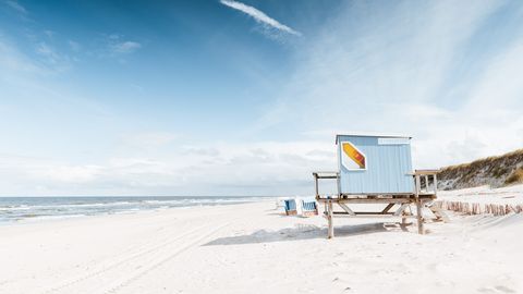 Ein blauer Rettungsschwimmerstand steht auf einem weiten, menschenleeren Sandstrand. Einige Strandkörbe sind in der Ferne sichtbar, während die Nordsee sich unter einem wolkenlosen Himmel erstreckt. Dünengras wächst im Hintergrund auf den Sandhügeln.