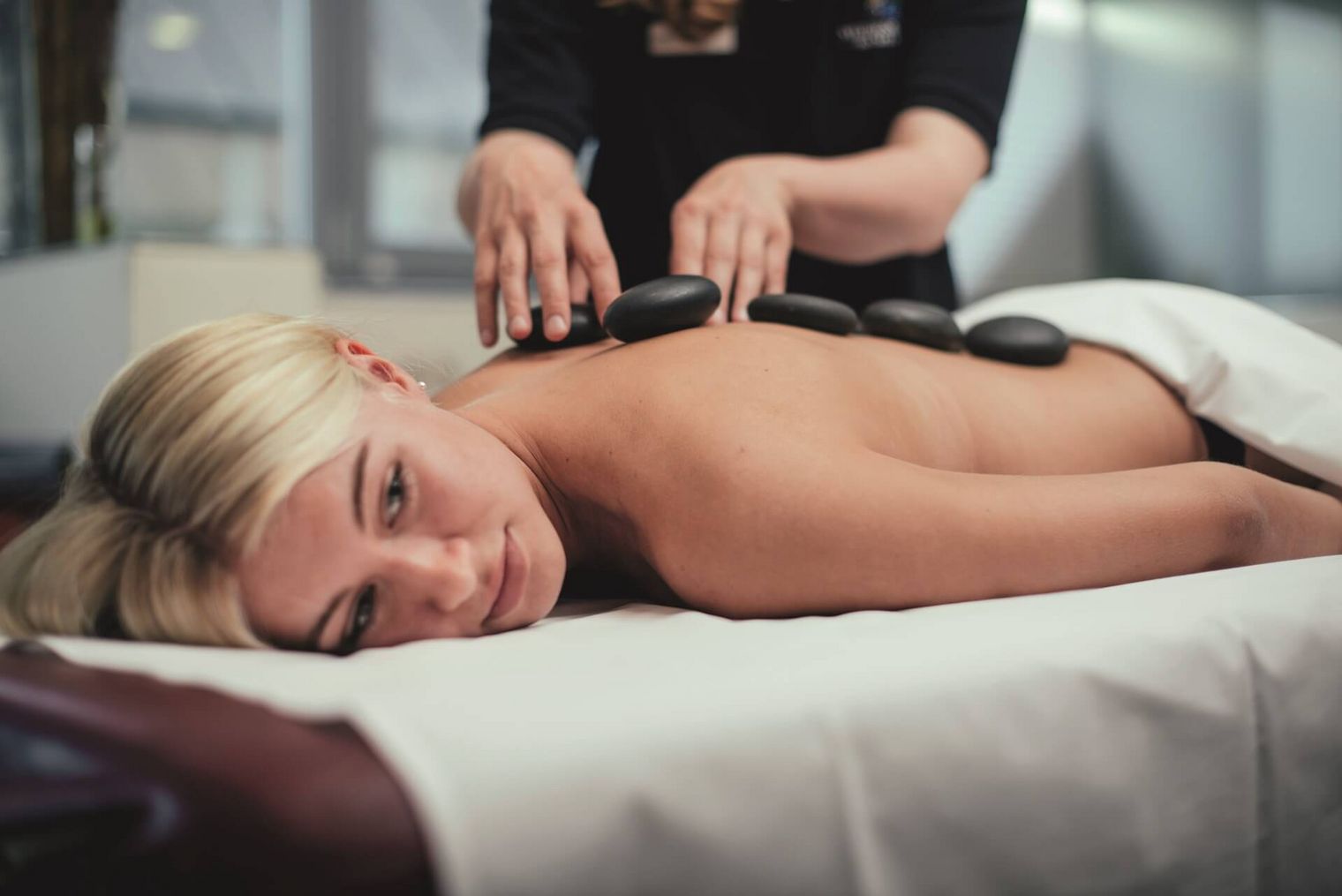 Eine blonde Frau erhält eine Hot-Stone-Massage auf einer Massageliege in einem hellen Wellnessraum.