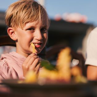 Ein Kind in einem rosa Pullover isst Pommes, während es an einem Tisch im Freien sitzt.