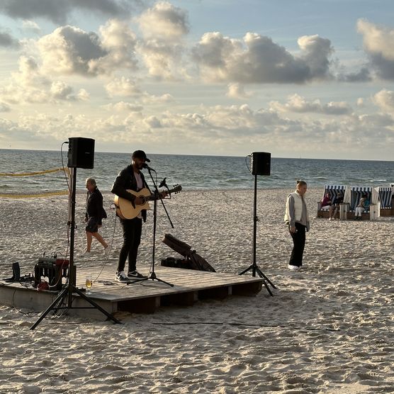 Ein Musiker spielt Gitarre und singt auf einer kleinen Bühne am Strand, umgeben von Lautsprechern. Im Hintergrund sind das Meer, der Himmel mit Wolken und einige Strandkörbe zu sehen.