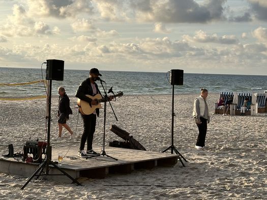 Ein Musiker spielt Gitarre und singt auf einer kleinen Bühne am Strand, umgeben von Lautsprechern. Im Hintergrund sind das Meer, der Himmel mit Wolken und einige Strandkörbe zu sehen.